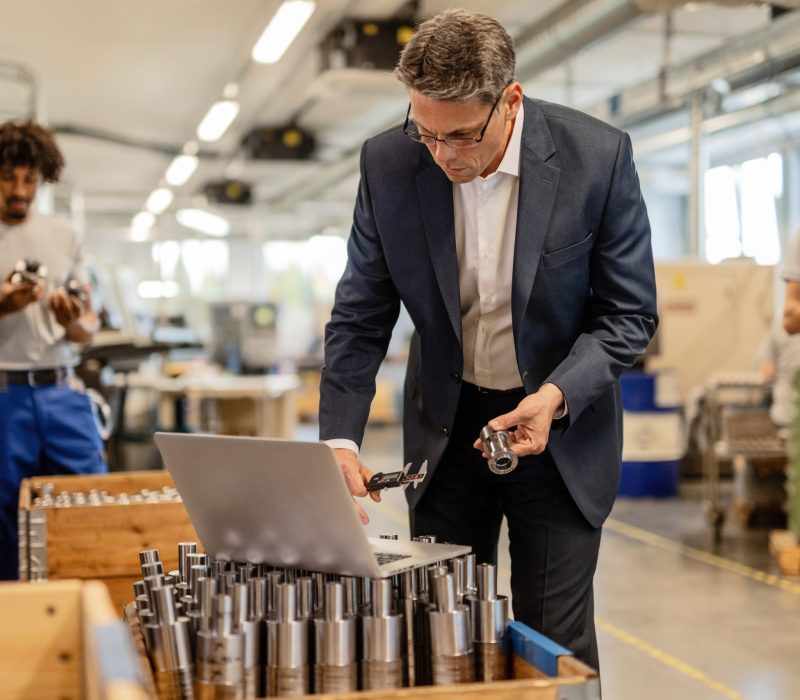 Factory manager reading instructions on a computer while checking measurements of steel bolt in industrial building. Manual workers are in the background.